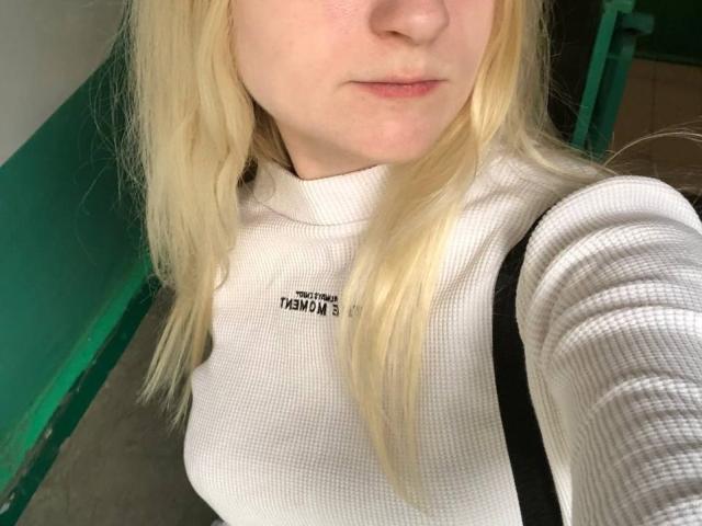 HyacinthBellin has blonde hair, light skin, and is wearing a white ribbed turtleneck that shows her small tits. She's holding a black strap with a green background.
