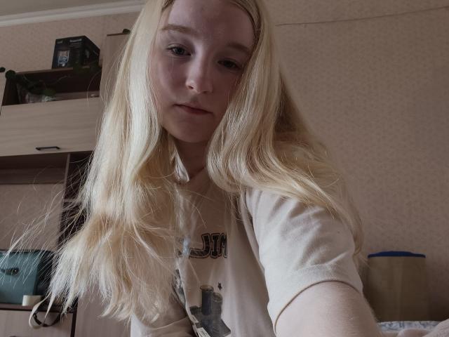 Blond-haired HyacinthBellin with pale skin and light freckles, wearing a beige shirt, taking a selfie in a dimly lit room, showing a relaxed, slightly curious expression.