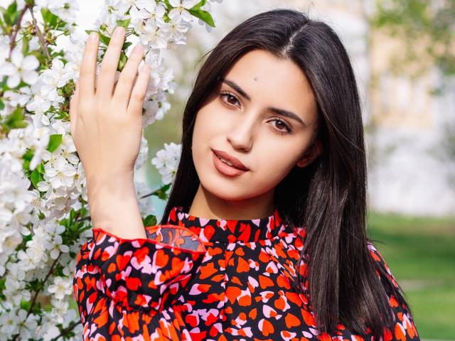 JasmineDay with dark hair, brown eyes, and light skin, wearing a red and black heart-patterned dress, touching white flowers with her right hand, smiling gently.