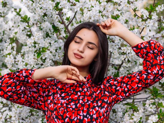 JasmineDay with dark hair, closed eyes, and red patterned shirt, arms raised, surrounded by white blossoms, looking relaxed and happy, showing her tits subtly through her shirt.