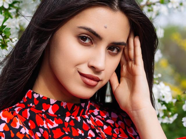 JasmineDay with dark brown hair, brown eyes, and a small dot on her forehead, wearing a red and black heart-patterned dress, touching her face with a slight smile.