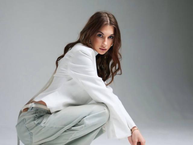 KatherinaEdnie has wavy brown hair, wearing a white long-sleeve shirt and light green pants, squatting with a subtle smile, showing a small butt and her lower back.