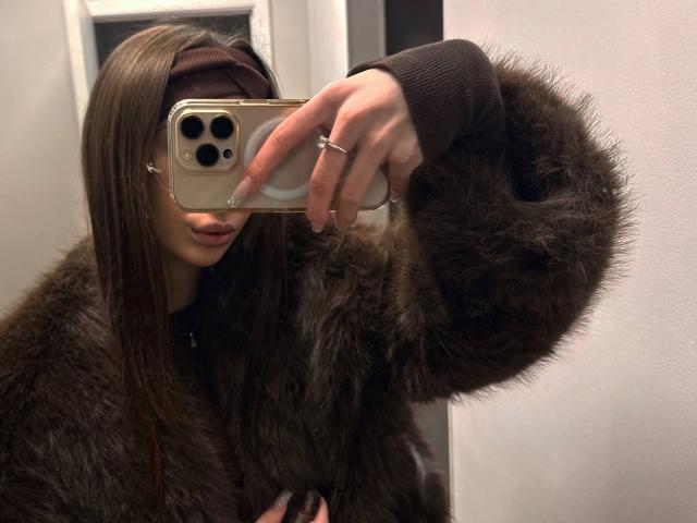 KatherinaEdnie in a brown fur coat, taking a mirror selfie with a gold iPhone, visible ring on her finger, long brown hair, and a brown headband.