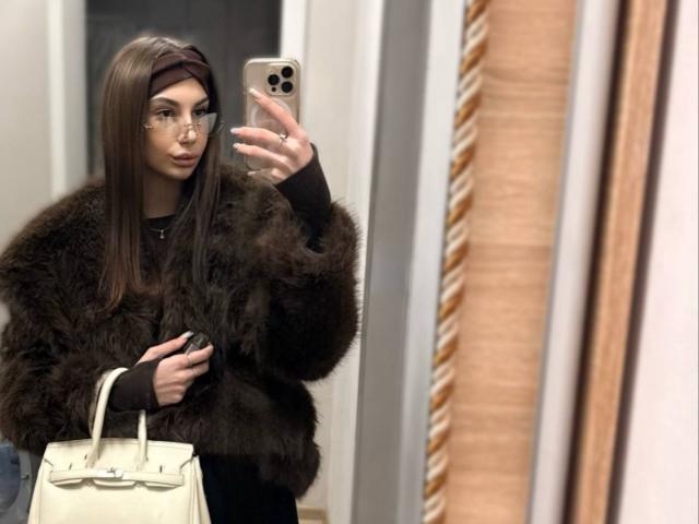 KatherinaEdnie with long brown hair, glasses, and a fluffy dark brown fur coat, holding a white handbag, taking a mirror selfie, looking at the phone.
