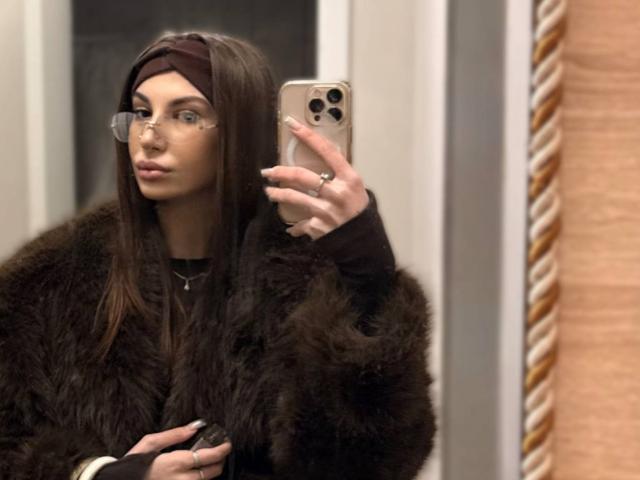 KatherinaEdnie with long brown hair, glasses, and brown headscarf, wearing a fluffy dark brown coat, taking a mirror selfie with an iPhone, showing a ring on her finger.