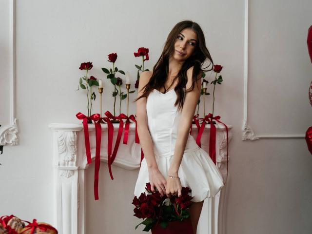 KatherinaEdnie with long brown hair, wearing a white dress, holding red roses, standing in front of a white mantel with red ribbon and more red roses.