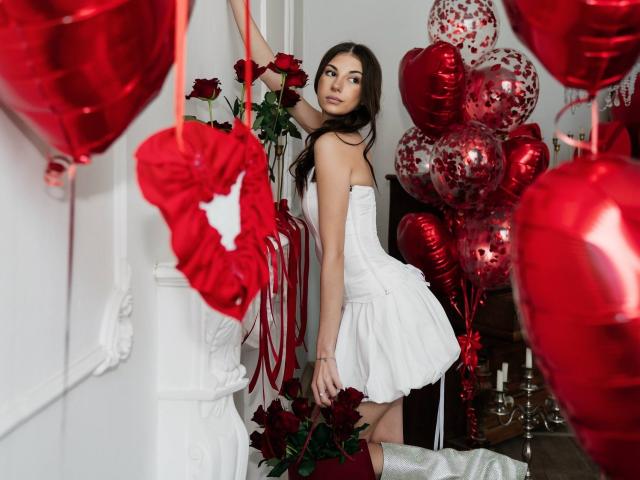 KatherinaEdnie with red rose, white strapless dress, and white boots, standing in room filled with red heart balloons and red roses, looking back with a slight smile.