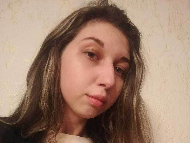 Sanella has light brown skin, long brown hair with blonde highlights, and is looking slightly to the side with a neutral expression. She's wearing a black top.
