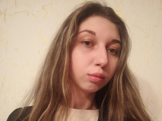 Sanella has long, messy brown hair, slightly puffy pink lips, and a neutral expression. She's wearing a black and white top, with light skin and brown eyes.