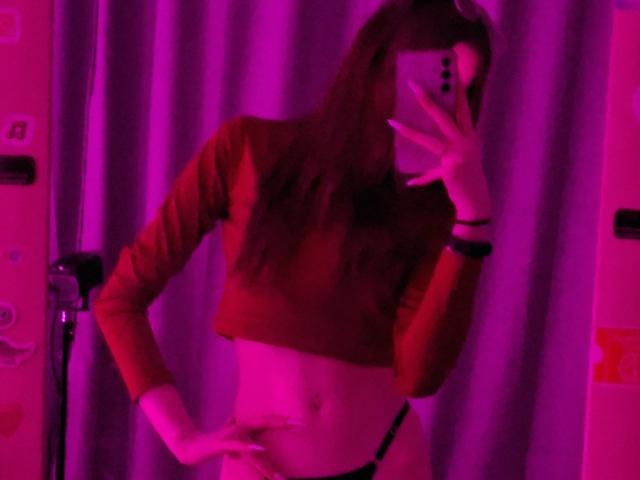 LeticiaEttl with long brown hair, red crop top, and black thong, holding phone in front of face, pink-purple lighting, hand on hip, mirror selfie, slim with small tits.
