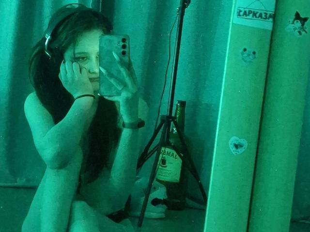 Nude LeticiaEttl with headphones, holding phone, mirror selfie, green-tinted. Bottle on tripod, long dark hair, slight smile, hands near face, green room, visible ass, mirror reflection.