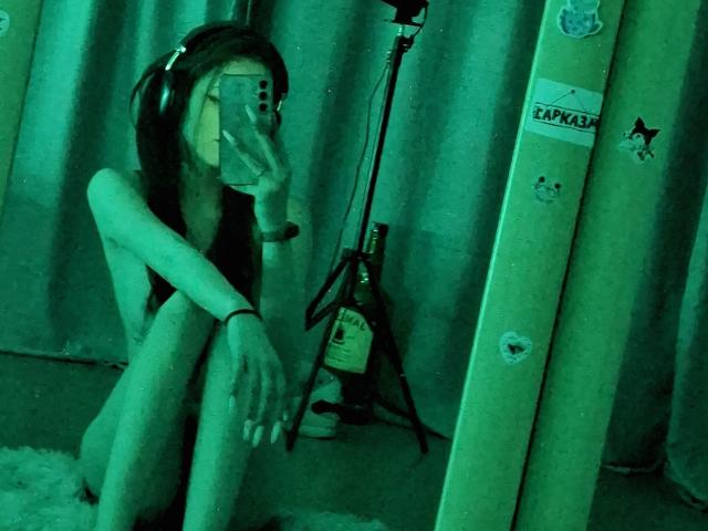 LeticiaEttl sits naked in green light, legs crossed, wearing headphones, holding phone, reflection in mirror, standing microphone, and ski boots in background, showing slim, pale body, small tits.