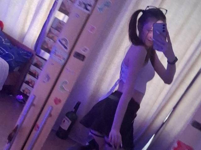 LeticiaEttl with pigtails, white crop top, black skirt, holding phone, mirror selfie, blue light, bottle on floor, reflection, smiling, glasses, looking back, medium tits, cute.
