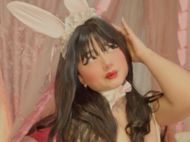 AnnieGrey, con i capelli neri, orecchie da coniglio bianche e fard rosa, che si tocca la testa, indossa una collana rosa e rossetto rosso, mostrando il suo seno prosperoso, di colore rosa..