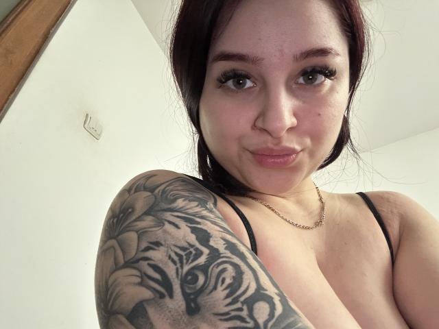 LucieRed has a tattooed arm with a tiger face, medium tits, and a cute smile. She's wearing a black strap and a gold necklace. Red hair, brown eyes.