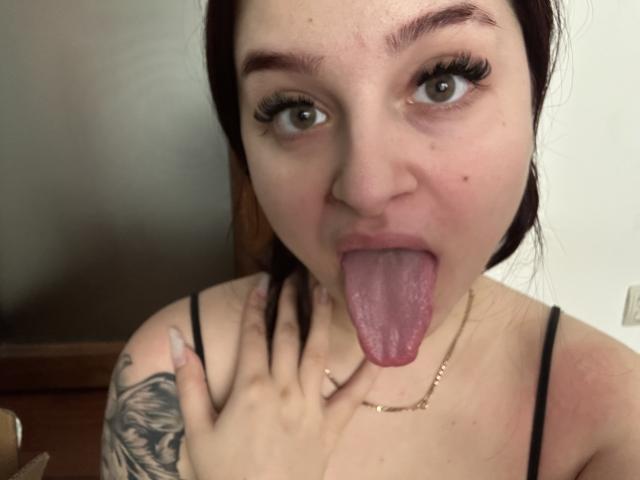 LucieRed with tongue out, blue eyes, black eyebrows, long eyelashes, black hair, tattoo on right arm, black spaghetti strap top, hand near mouth, necklace, looking at camera.