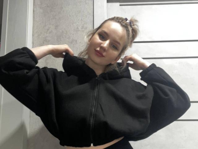 ChereServais, con cabello rubio recogido en un moño despeinado, vestida con una sudadera negra con cremallera, ajustándose el cabello con ambas manos, sonriendo ligeramente, mostrando sus pechos..