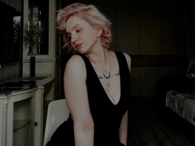 ChereServais con cabello rosa, vestido negro de corte profundo en V, mostrando los senos, tatuaje sobre el pecho, ligeramente inclinada, mirando hacia abajo, sentada en una habitación oscura..