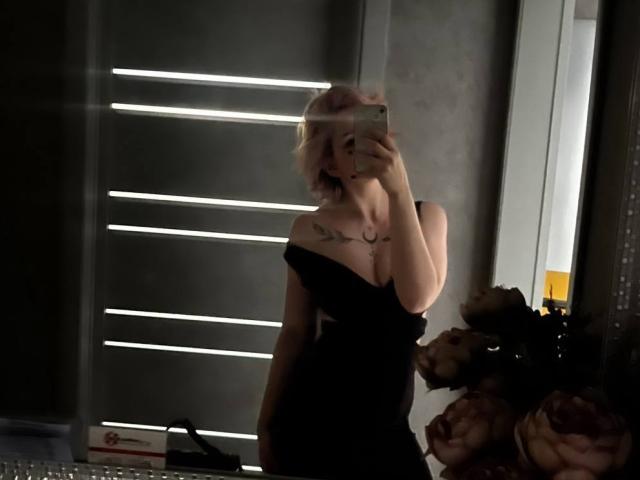 ChereServais con cabello rosa, vestido negro con un hombro descubierto, sosteniendo un teléfono en el espejo, mostrando los senos, tatuaje en el pecho, en una habitación con poca luz, arreglo floral a la derecha..
