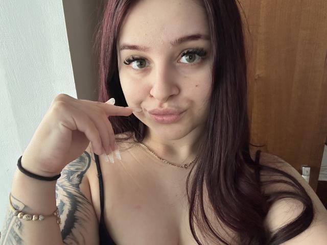 LucieRed with long red hair, green eyes, and tattoos on her right arm, pouting with her finger on her cheek, wearing a black top and a gold necklace.