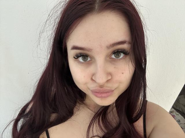 LucieRed has pale skin, green eyes, and long red hair. She's making a pouty face with full lips, wearing a black strap top. Her tits are small and cute.