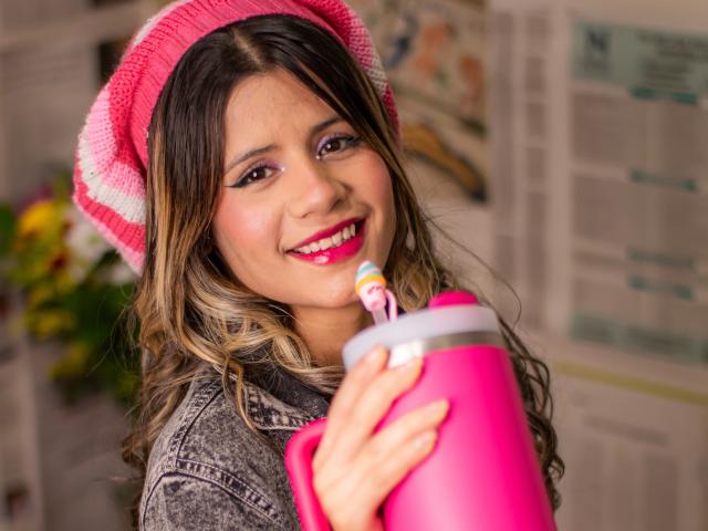 SheylaSpooner with curly brown hair, pink beanie, red lipstick, holding pink thermos with striped straw, smiling, wearing gray denim jacket, in colorful indoor setting.
