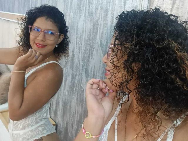 VioletJoness, con cabello castaño rizado, gafas, lencería blanca de encaje, lápiz labial rojo, sonriendo, mirando en el espejo, mano derecha cerca de la barbilla, mano izquierda sobre la encimera, pulsera rosa, joyería discreta..