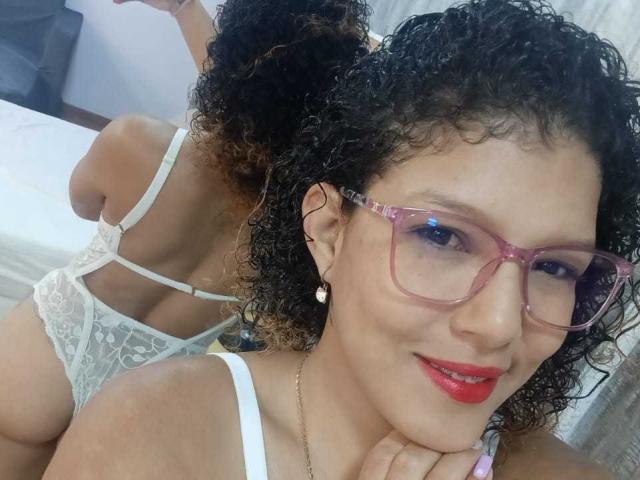 VioletJoness with curly hair, pink glasses, red lipstick, white lace lingerie, showing her back and butt, smiling at the camera, in a mirror selfie.