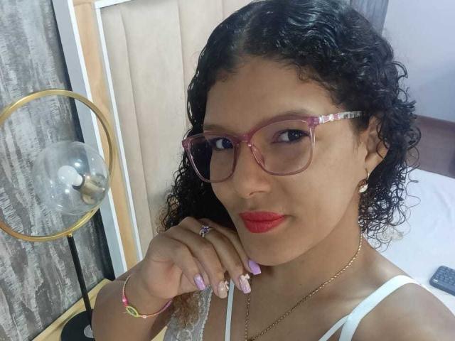 VioletJoness, con cabello negro rizado, gafas rosas, lápiz labial rojo, blusa blanca, uñas rosas, anillo, pulsera y collar, posando con el dedo cerca de la boca, mostrando sus senos..