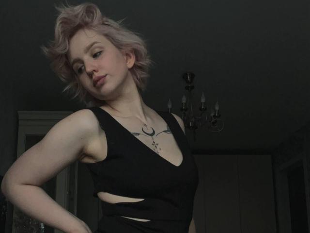 ChereServais, con cabello rosa corto, vestido negro con aberturas, pechos pequeños, tatuaje en el pecho, de pie en una habitación oscura, mirando hacia abajo, con los brazos ligeramente levantados, un candelabro en el fondo..
