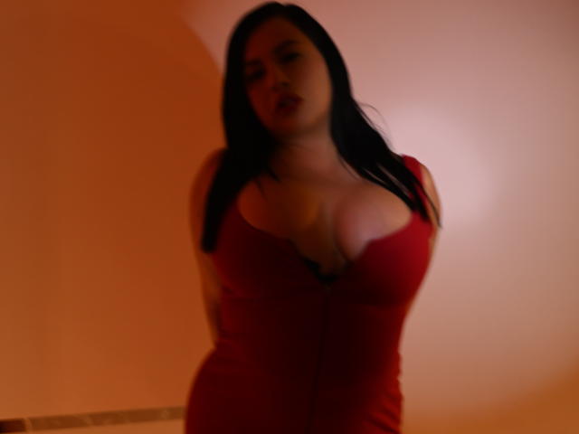 NahomiRosse has large, prominent tits in a tight, red dress with a deep cleavage, dark hair, and a slightly blurred, sexy, sultry face. Warm, orange lighting.