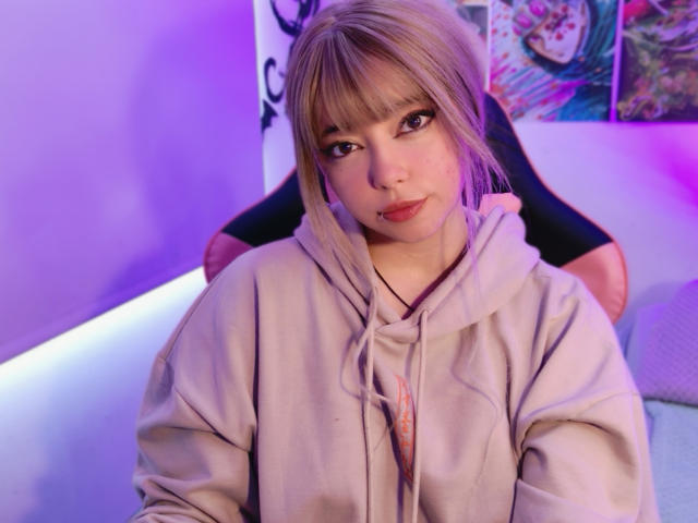 Arieyaara with blonde hair, bangs, and red lipstick, wearing a beige hoodie, sitting with a black chair behind her, purple and pink lighting, looking at the camera.