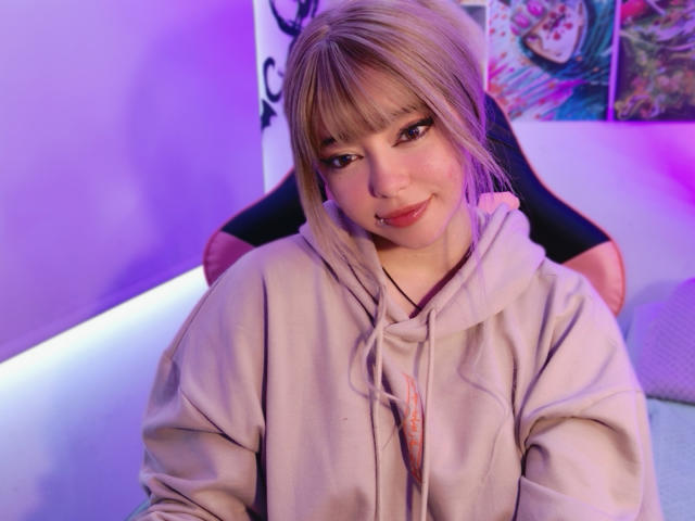 Arieyaara, com cabelo loiro, franja e um sorriso adorável, vestindo um moletom bege, sentada com as pernas levantadas, e uma calcinha preta à mostra, em um quarto com iluminação roxa..