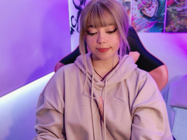 Arieyaara with blonde bangs, wearing a beige hoodie, sitting, eyes down, black gloves, purple and pink lighting, small nose piercing, cute, soft smile, slight blush, relaxed.