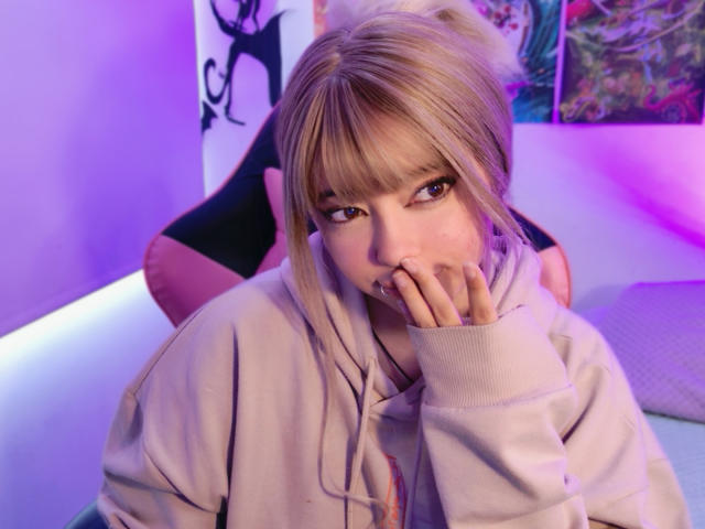 Arieyaara with blonde hair, bangs, and brown eyes, wearing a beige hoodie, has her hand over her mouth, sitting with a purple and pink-lit background.