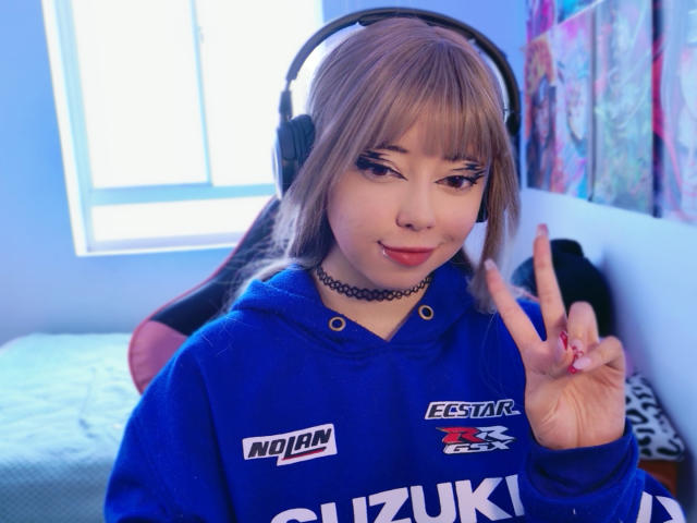 Arieyaara with blonde hair, wearing a blue hoodie, black choker, and headphones, flashing a peace sign, has a cute face, and is looking at the camera.