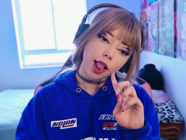 Arieyaara with light brown hair, blue hoodie, black choker, and headphones, sticking out tongue with finger touching it, pink nails, in a brightly lit room.