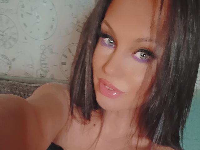 A MadisonTayler69 tem sombra roxa brilhante, lábios rosa e brilhantes e cabelo preto e liso. Seus olhos azuis estão olhando para a câmera com um leve sorriso. Ela está mostrando seus seios..