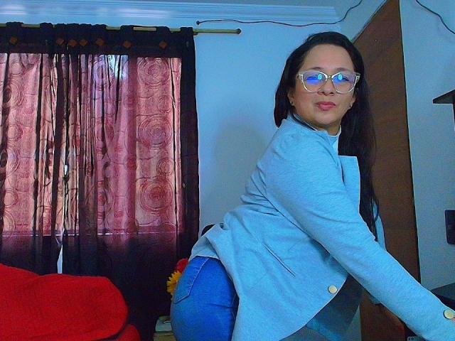 VictoriaRousse with glasses, blue jacket, and blue jeans, bending forward, showing her ass, red couch in background, dark curtains with pink pattern, long black hair, smiling.