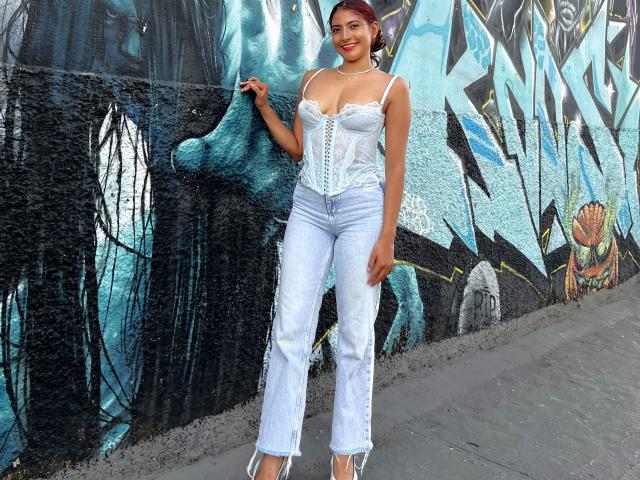 MiaBianchi has red hair, white lace bustier, showing her tits, and blue jeans, standing against a graffiti wall, wearing white heels, smiling with one hand on her hip.