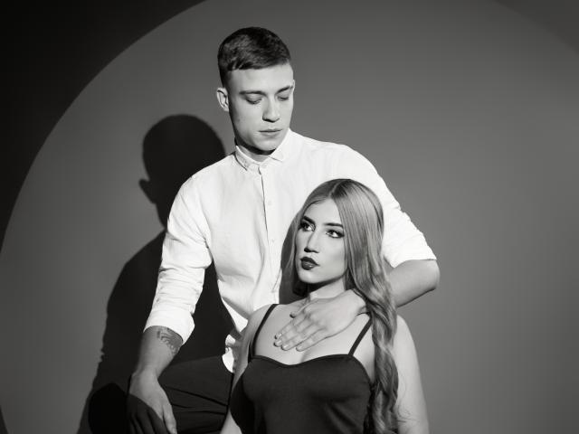 SecretLoversVIP con i capelli corti e la camicia bianca, con una mano sulle tette di SecretLoversVIP con i capelli lunghi, vestito scuro e trucco occhi intenso, in piedi contro un muro spoglio..