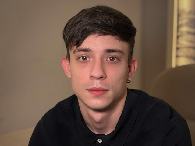 SecretLoversVIP has a cute face, short brown hair, brown eyes, and a small earring in his left ear. He's wearing a black shirt and has a neutral expression.