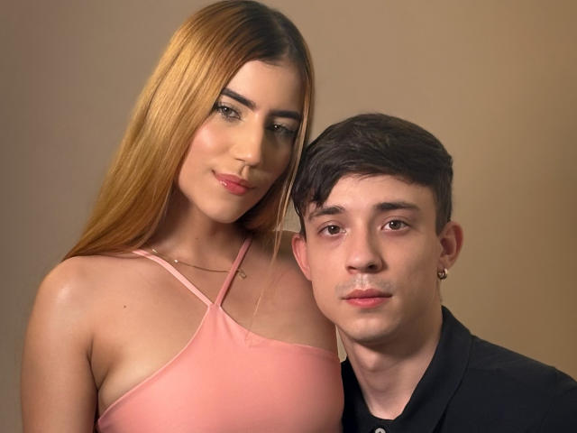SecretLoversVIP with long brown hair and pink top, showing her tits, stands next to a guy with short brown hair and black shirt, both with subtle smiles.