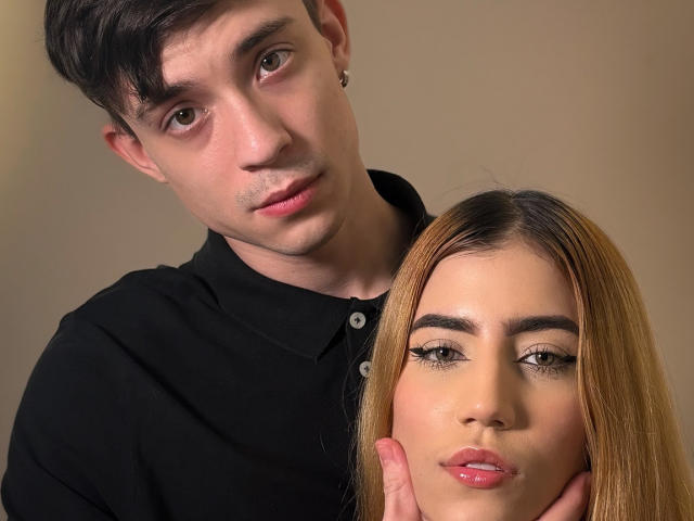 SecretLoversVIP avec des cheveux bruns courts et une boucle d'oreille, un polo noir, tenant une autre SecretLoversVIP avec de longs cheveux blonds, du rouge à lèvres rose et un eye-liner ailé, sur un fond beige..