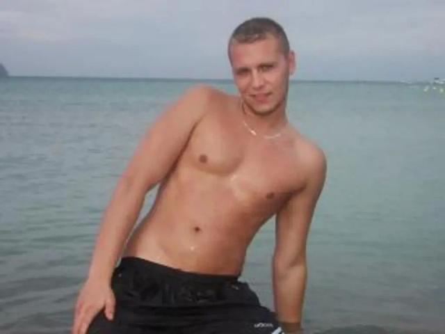 Homme torse nu en feu - bronzé, avec des cheveux bruns courts, une petite poitrine, portant un short noir, debout dans de l'eau peu profonde, légèrement penché vers la droite, souriant..