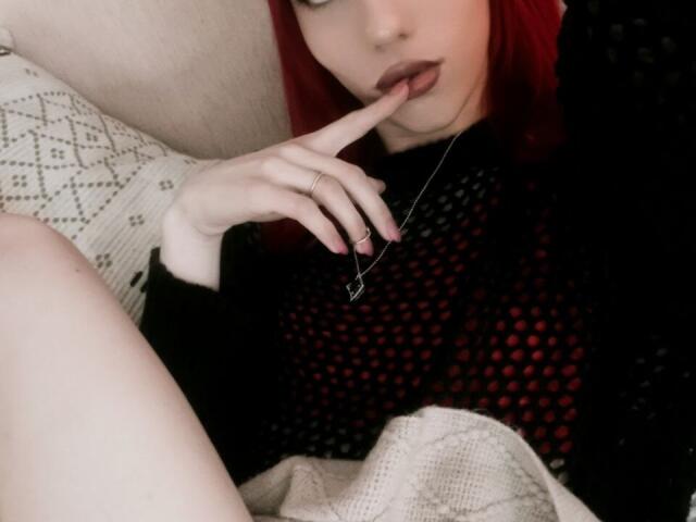 KiranaHayes with red hair, finger in mouth, black mesh top, white knee sock, and ring, showing off her pale skin and delicate fingers, lying on a beige sofa.
