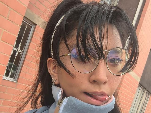 VioletXBloom's face with black puffy hair, gold-rimmed glasses, and a silver lip piercing. She's wearing a blue zip-up and has multiple ear piercings. She's sticking out her tongue.