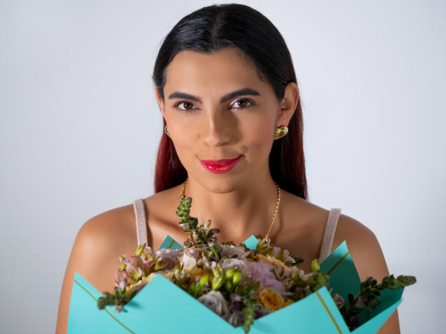 Carla Torres, con i capelli rosso scuro, rossetto rosso, orecchini dorati, top beige, che tiene una scatola verde con fiori rosa e bianchi, sorride leggermente e guarda la telecamera..