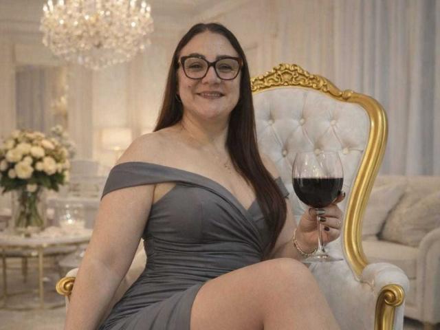LadyNylonOh with glasses, gray off-shoulder dress, holding wine glass with red wine, sitting on gold and white tufted chair, smiling, long brown hair, fair skin, in elegant room.