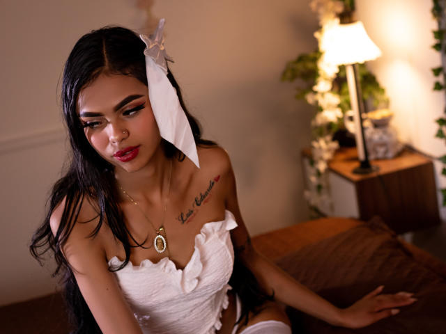 CrystalDavids with long black hair, white dress, and large white bow, showing small tits, tattoo on chest, red lipstick, and gold necklace, sitting on brown bed, looking down.